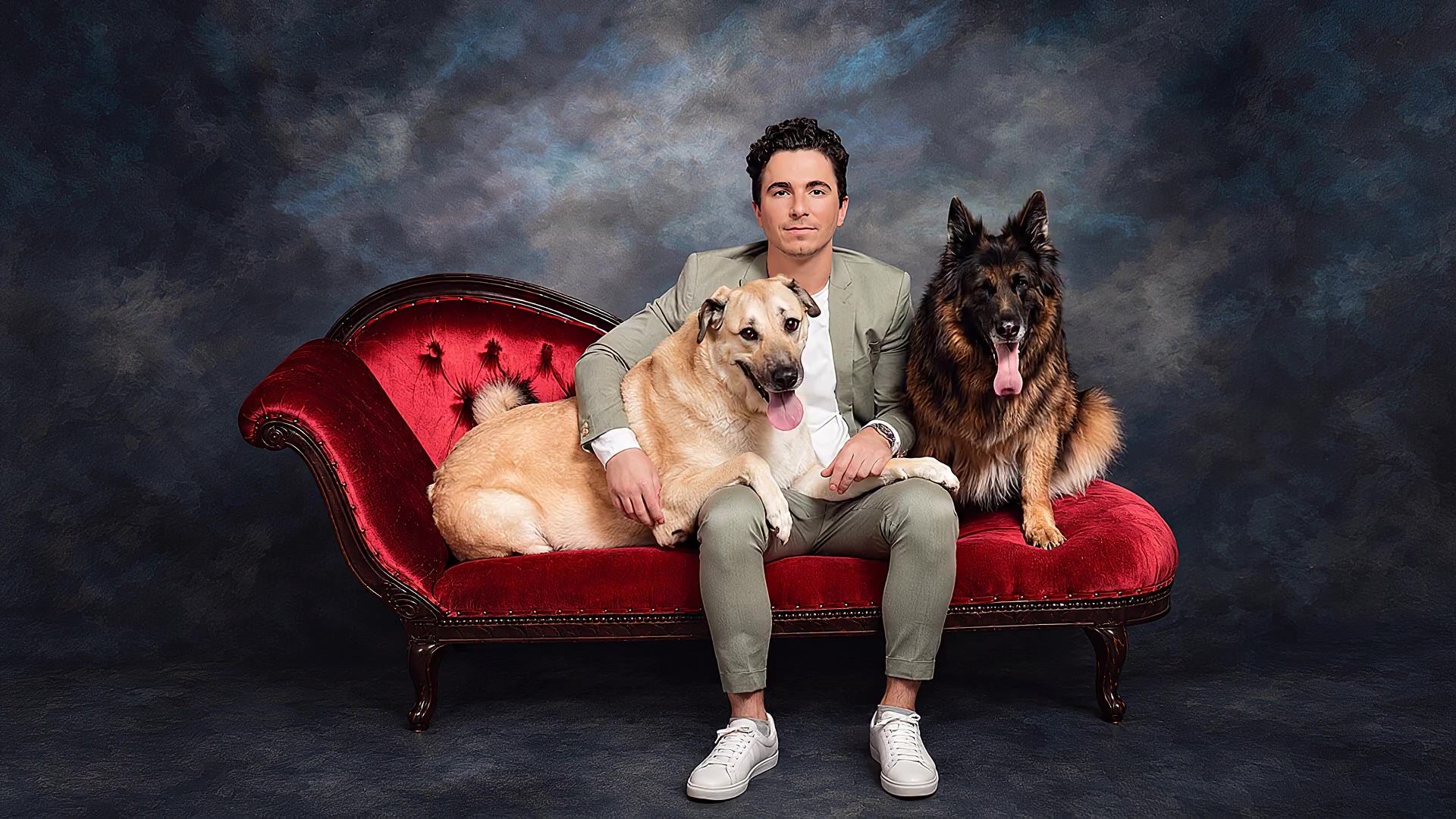 A man seated on a red sofa with two dogs against a dark studio backdrop.